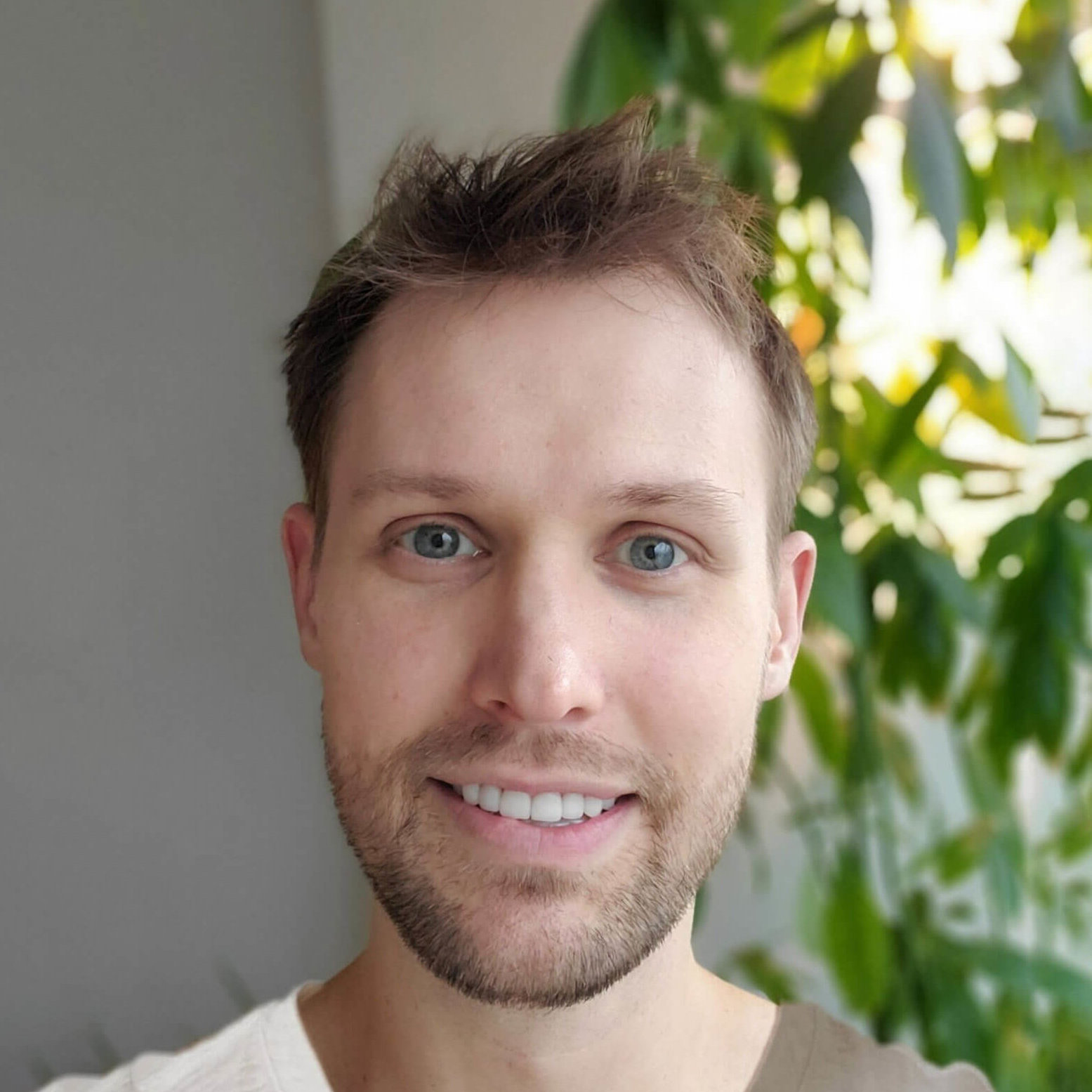 A photo of Scott Splunchic, who has short, dark blonde hair, short facial hair, and blue eyes. He is wearing a white shirt and is smiling pleasantly in a room with lush greenery behind him.